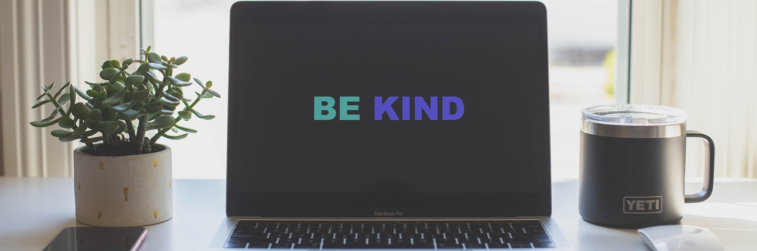 Header image for the BE K.I.N.D Embodied Wisdom Methodology. It's a laptop with the words BE KIND, with a small plant on the left and a coffee cup on the right.
